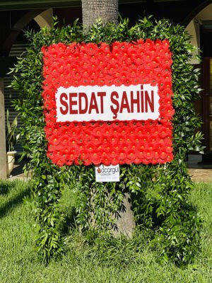 Adana açılış çiçek standı, yeşilliklerle kaplı ayaklı çerçeve içinde kırmızı çiçekli ve isimli kare pano.